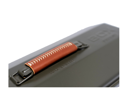 A close-up of a dark gray metal case with a brown leather handle wrap featuring white stitching and a black metal latch.