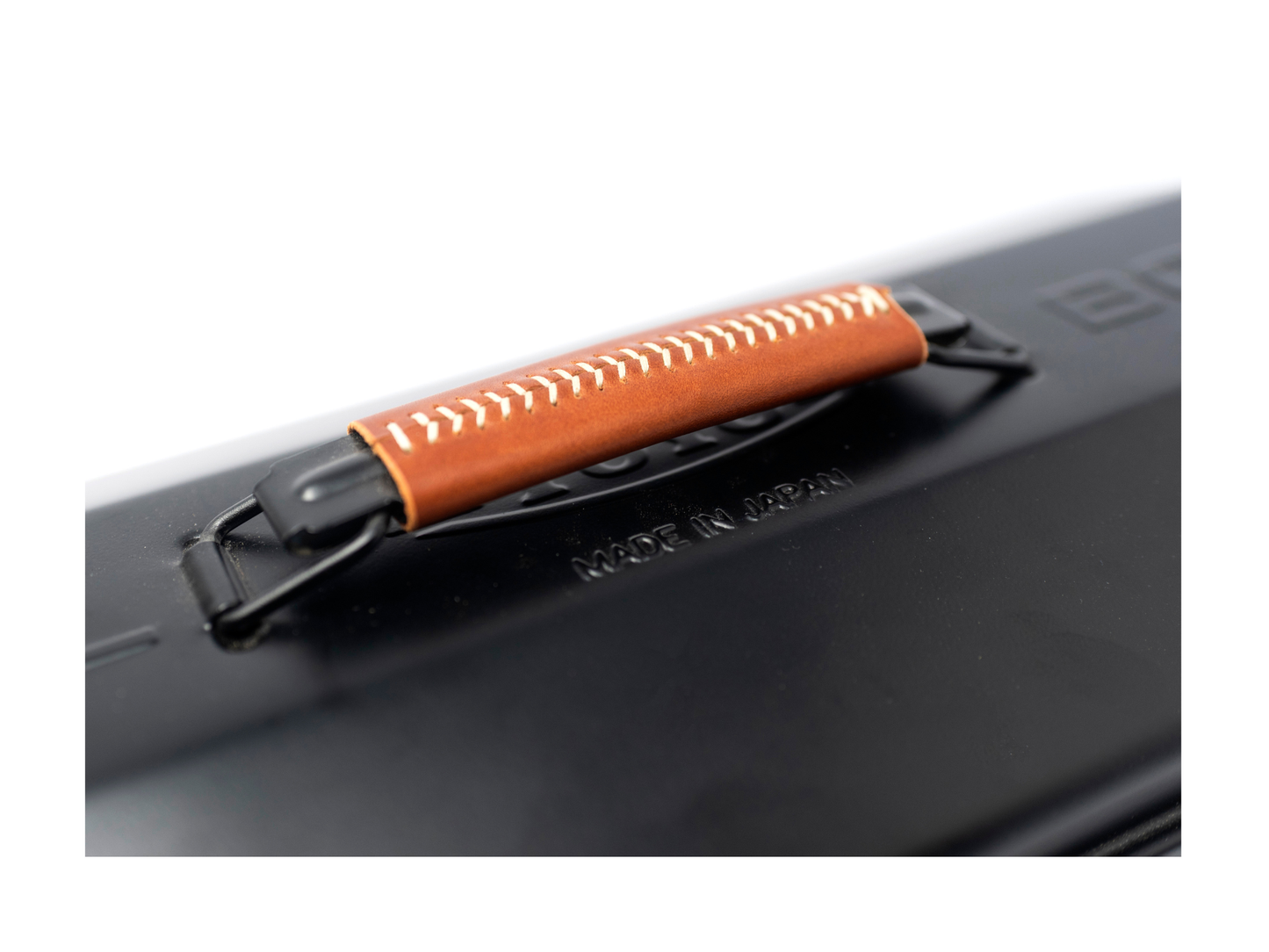 Close-up of a black hard case with a brown leather handle featuring white stitching and black metal hardware, on a white background.