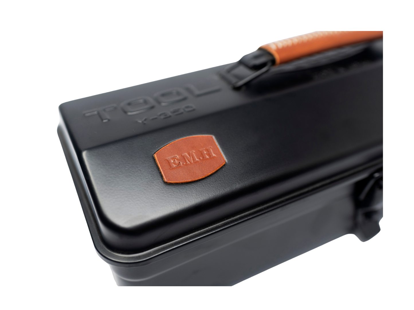 Close-up of a black metal tool case with an orange handle and an orange leather patch embossed with 'BMH'.