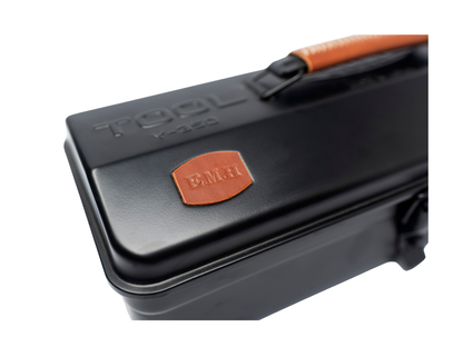 Close-up of a black metal tool case with an orange handle and an orange leather patch embossed with 'BMH'.